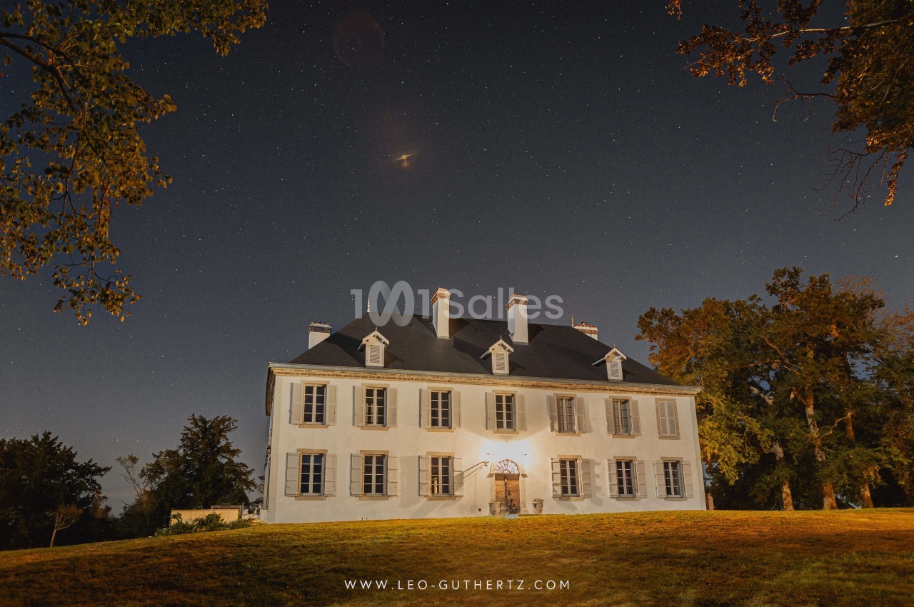 Façade d'un manoir blanc éclairé la nuit, entouré d'arbres sous un ciel étoilé.