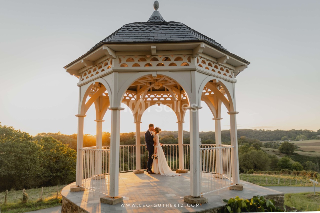 Un couple se tenant sous un kiosque en bois au coucher du soleil, entouré d'un paysage vallonné.