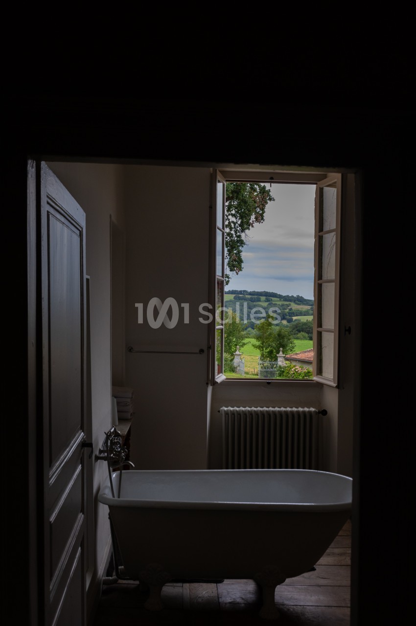 Salle de bain sombre avec une baignoire sur pieds, une fenêtre ouverte donnant sur un paysage verdoyant.