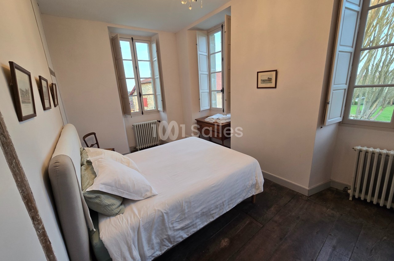 Chambre lumineuse avec lit double, parquet en bois sombre, murs clairs, fenêtres ouvertes et mobilier en bois.