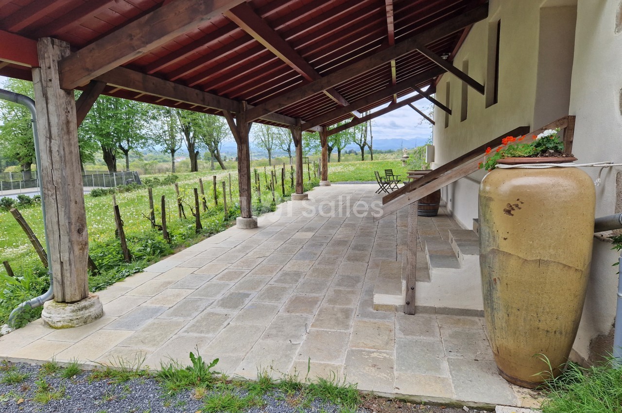Terrasse couverte en bois avec sol en pierre, grande jarre décorative et vue sur un jardin verdoyant.