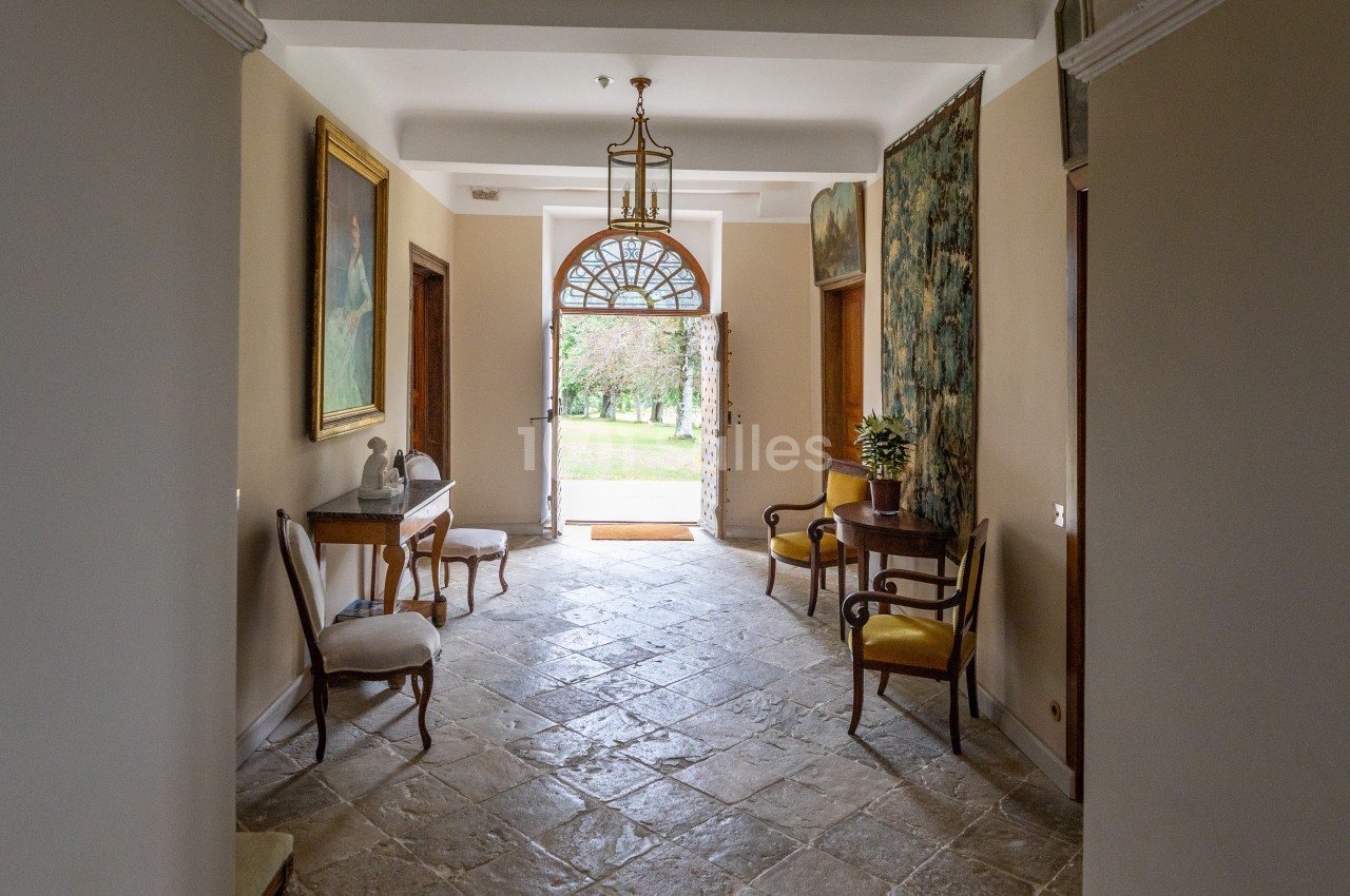 Couloir lumineux avec sol en pierre, mobilier ancien, tableaux aux murs et porte ouverte donnant sur un jardin verdoyant.