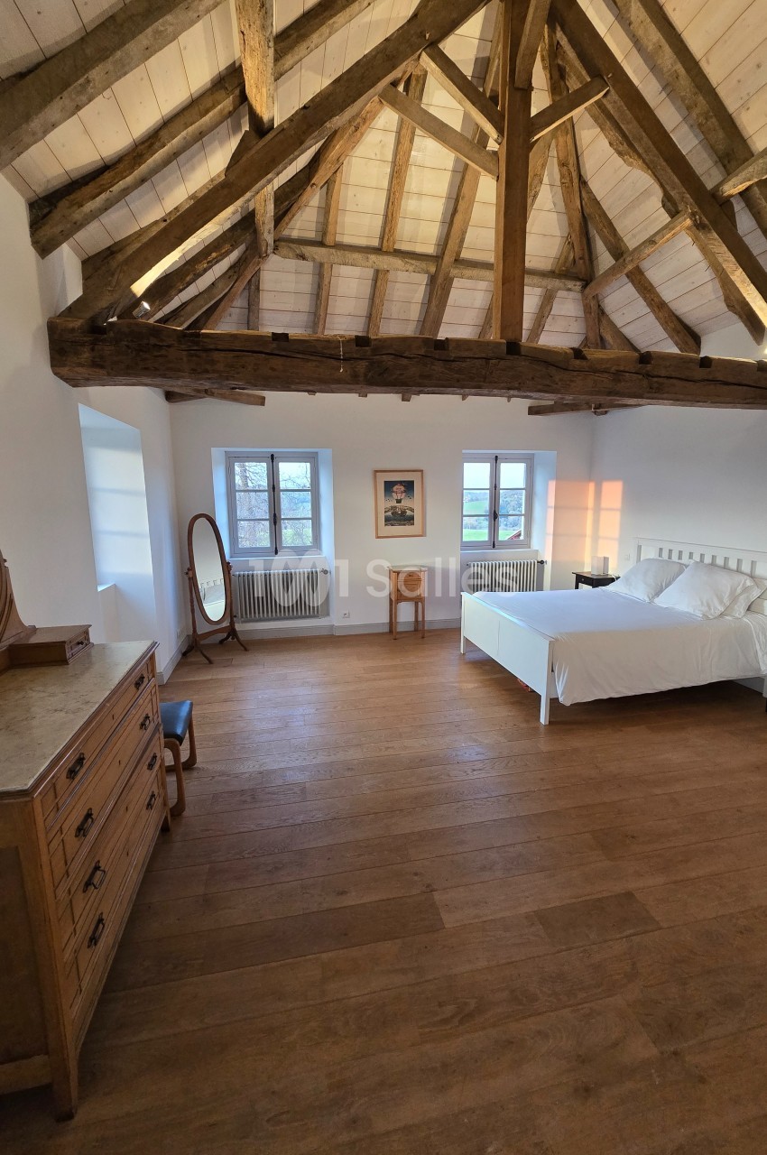 Chambre lumineuse avec poutres apparentes, lit double, commode en bois et fenêtres donnant sur l'extérieur.