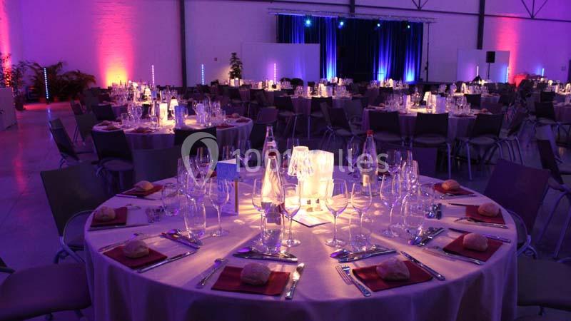 Salle de réception décorée avec des tables rondes dressées, éclairage coloré et ambiance festive.