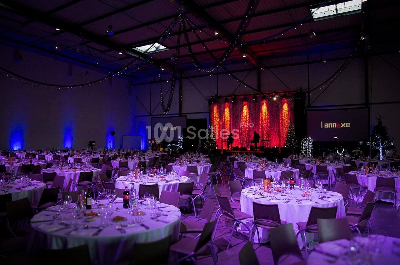 Salle de réception décorée avec des tables rondes dressées, éclairage tamisé et scène illuminée en arrière-plan.