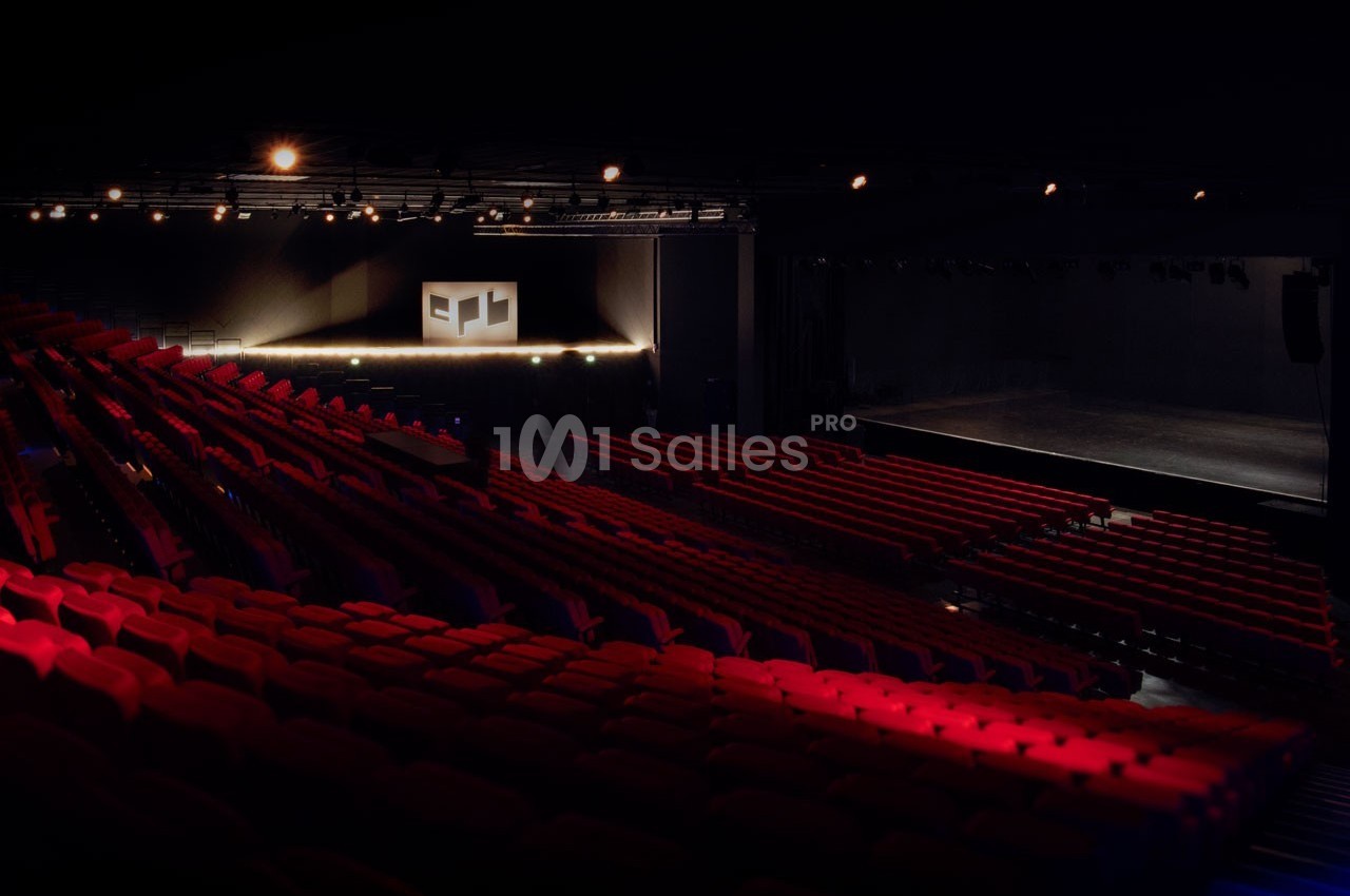 Salle de spectacle vide avec des rangées de sièges rouges et une scène éclairée en arrière-plan.