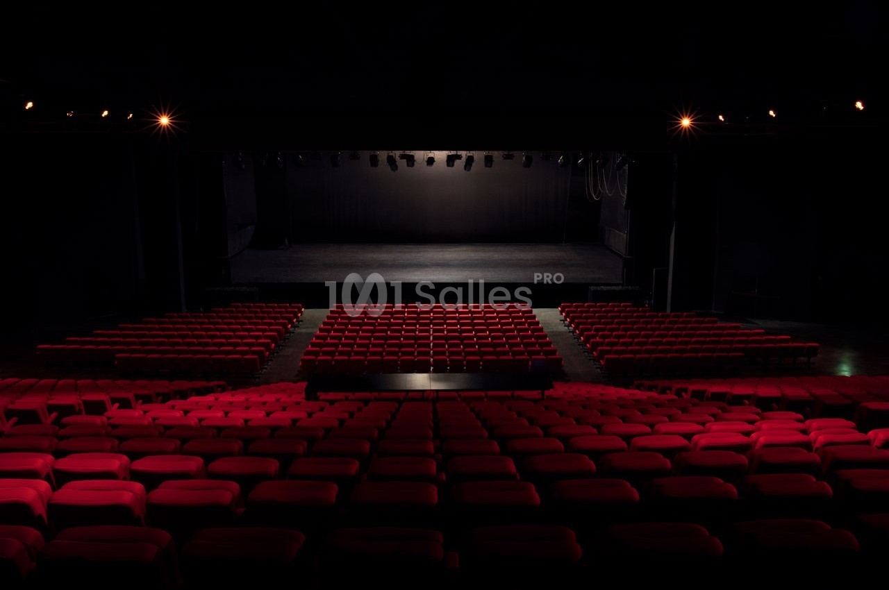 Salle de théâtre vide avec des rangées de sièges rouges et une scène éclairée en arrière-plan.