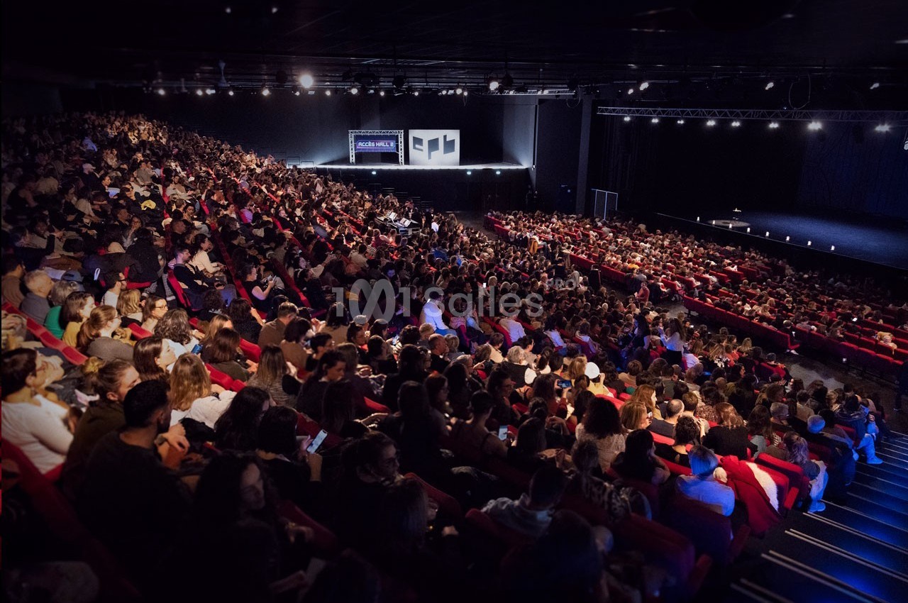 Salle de spectacle bondée avec des spectateurs assis sur des rangées de sièges rouges, face à une scène éclairée.