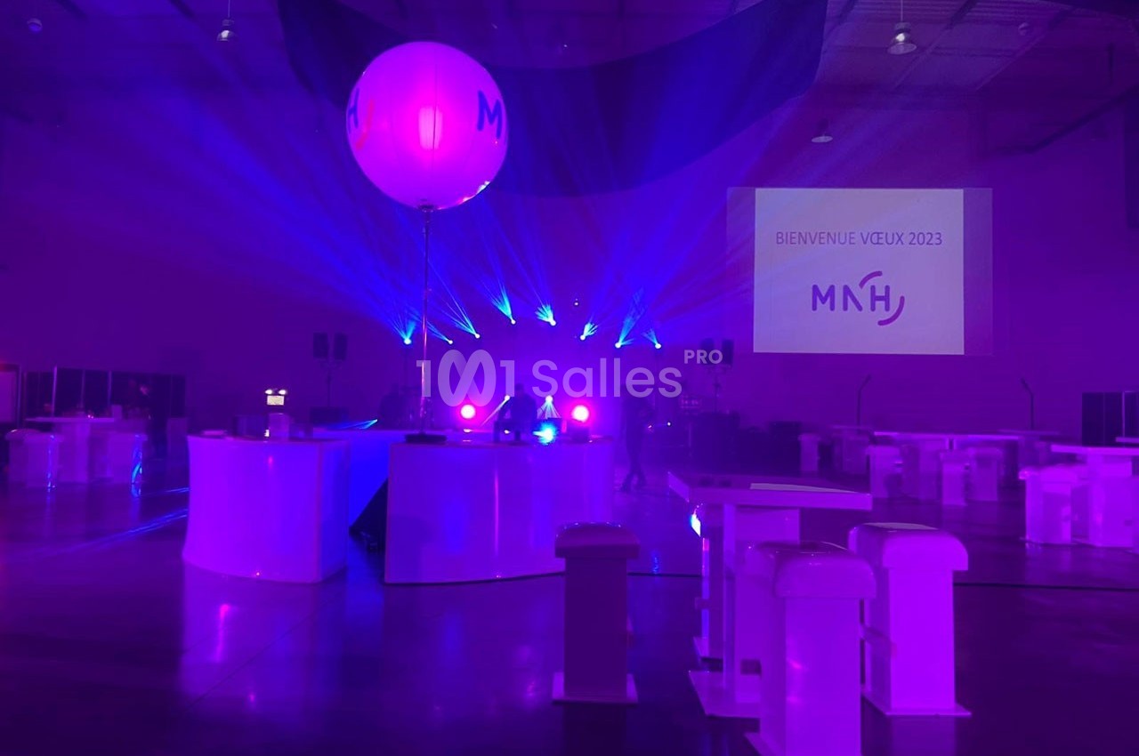 Salle événementielle éclairée par des lumières violettes, avec un ballon marqué ’MNH’ et un écran affichant ’Bienvenue Velix…