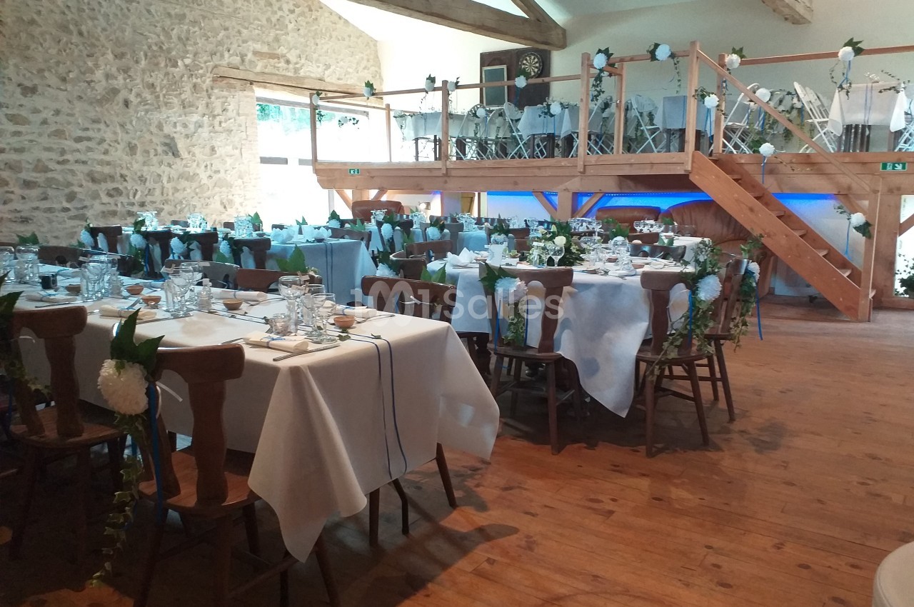 Salle de réception décorée pour un événement, avec tables dressées, chaises ornées et mezzanine en bois visible.