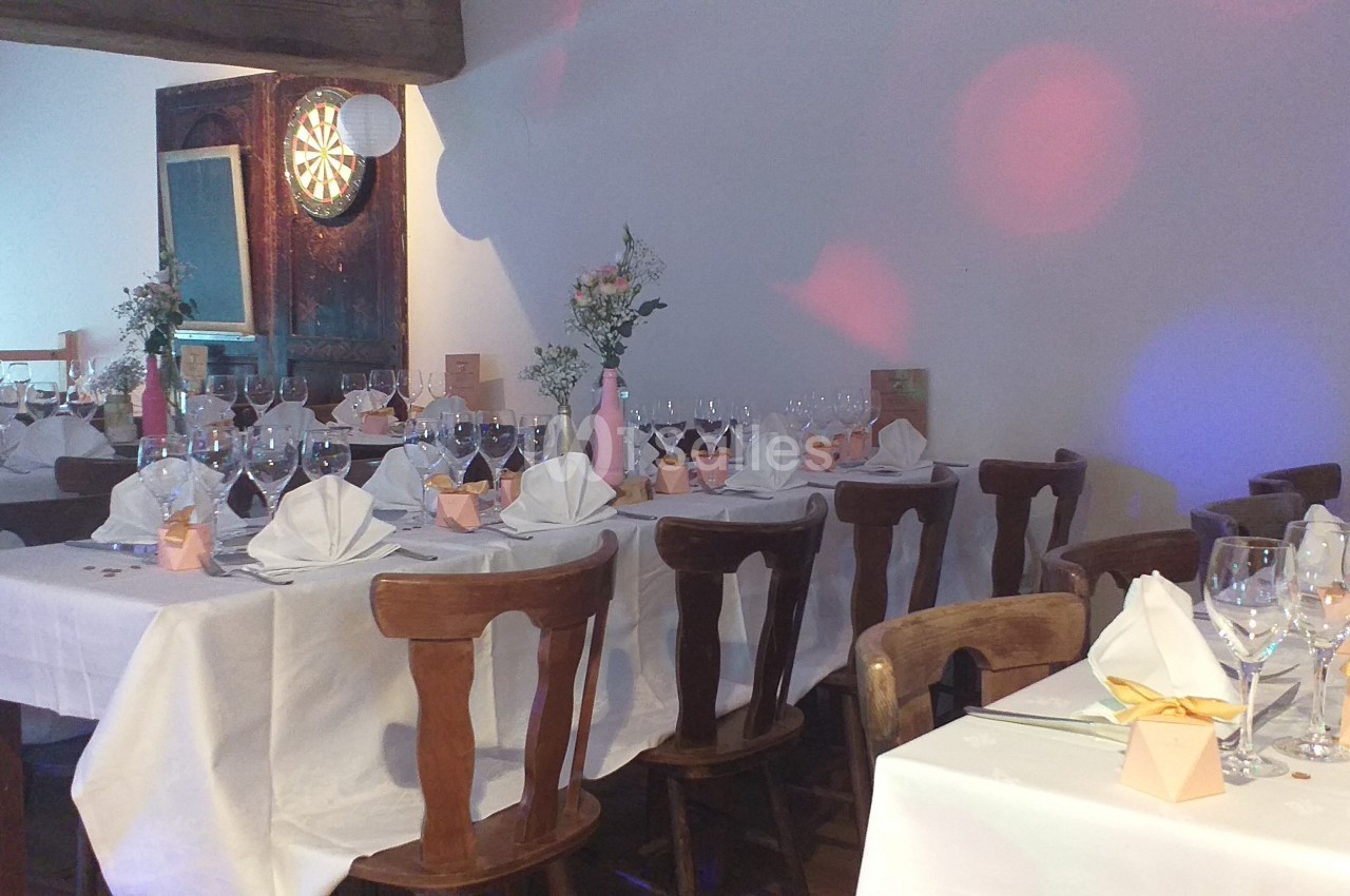 Salle décorée pour un repas festif avec tables dressées, nappes blanches, pliages de serviettes et arrangements floraux.