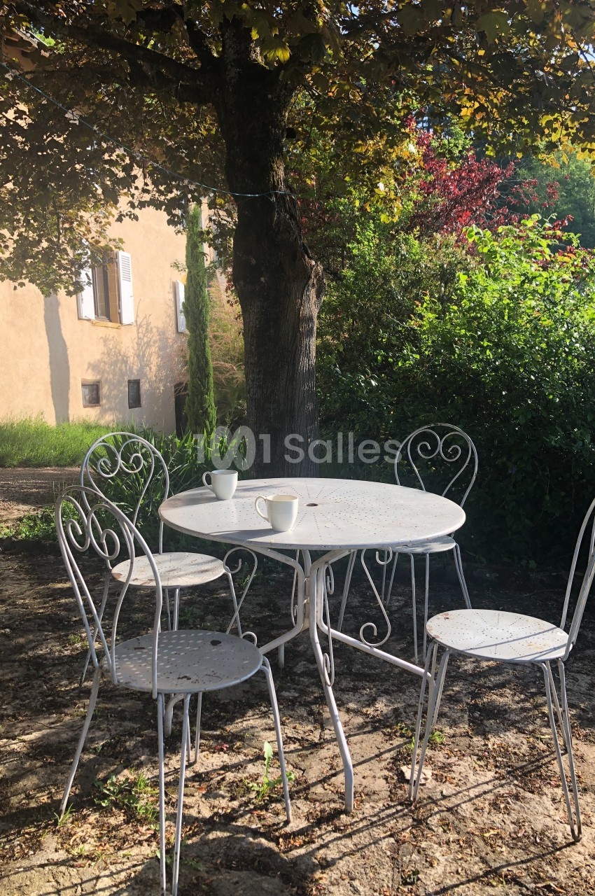 Table et chaises en métal blanc sous un arbre, avec deux tasses posées sur la table, dans un jardin ensoleillé.