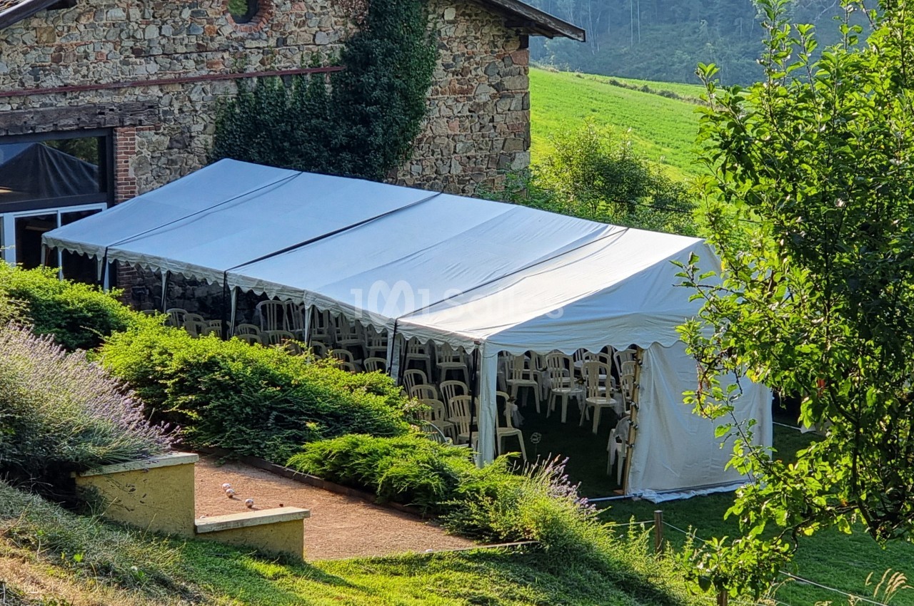 Tente blanche installée à côté d'une maison en pierre, entourée de verdure et de chaises visibles à l'intérieur.