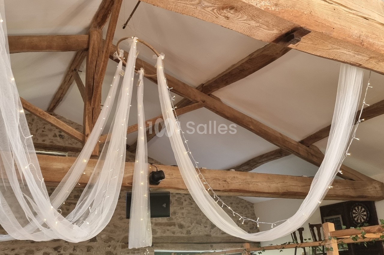 Plafond avec poutres en bois apparentes décorées de voilages blancs et de guirlandes lumineuses.