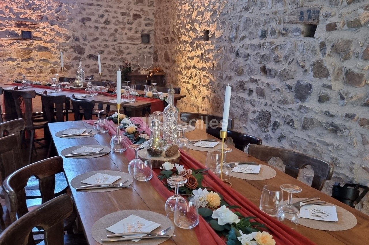 Table en bois décorée pour un repas festif, avec bougies, fleurs et vaisselle, dans une salle aux murs en pierre.