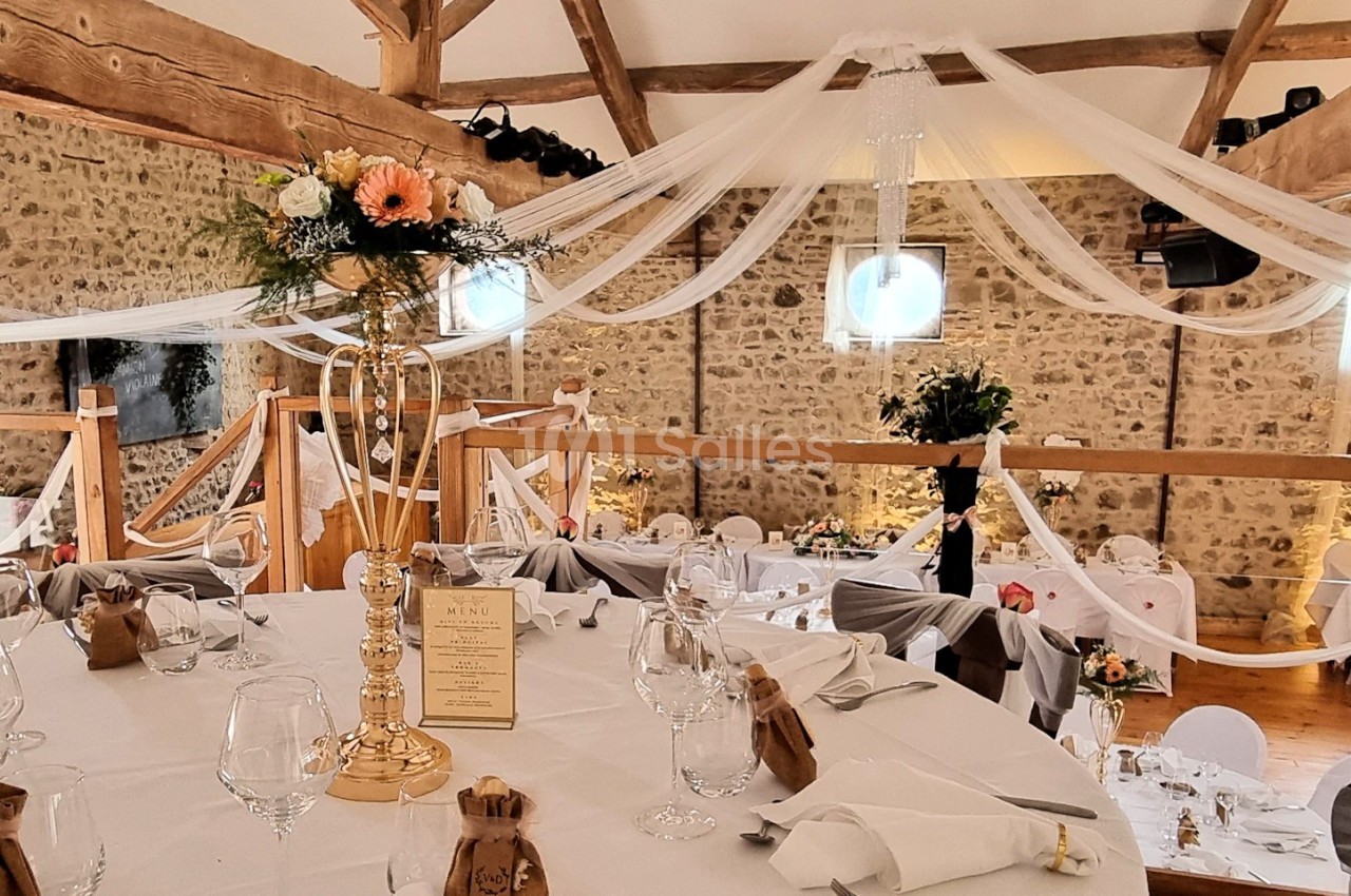 Salle de réception décorée avec des tables dressées, des fleurs et des voilages suspendus sous une charpente en bois.