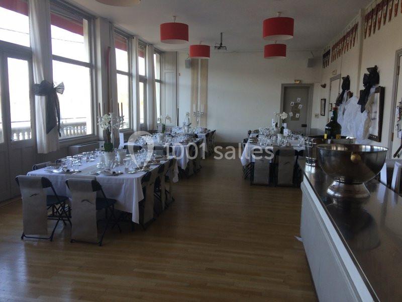 Salle de réception lumineuse avec tables dressées pour un événement, décorations sobres et grandes fenêtres.