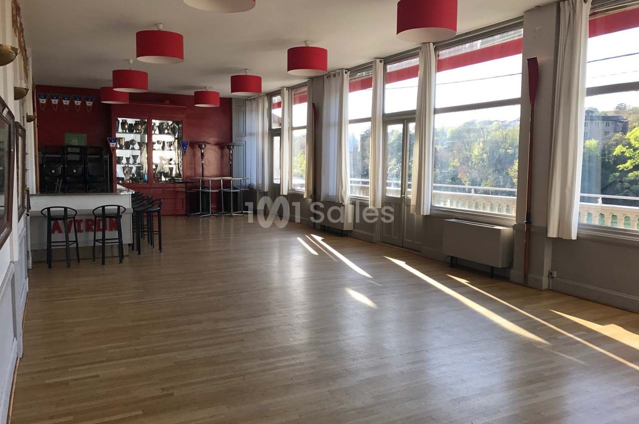Salle lumineuse avec parquet en bois, grandes fenêtres, rideaux blancs, murs rouges et trophées exposés.