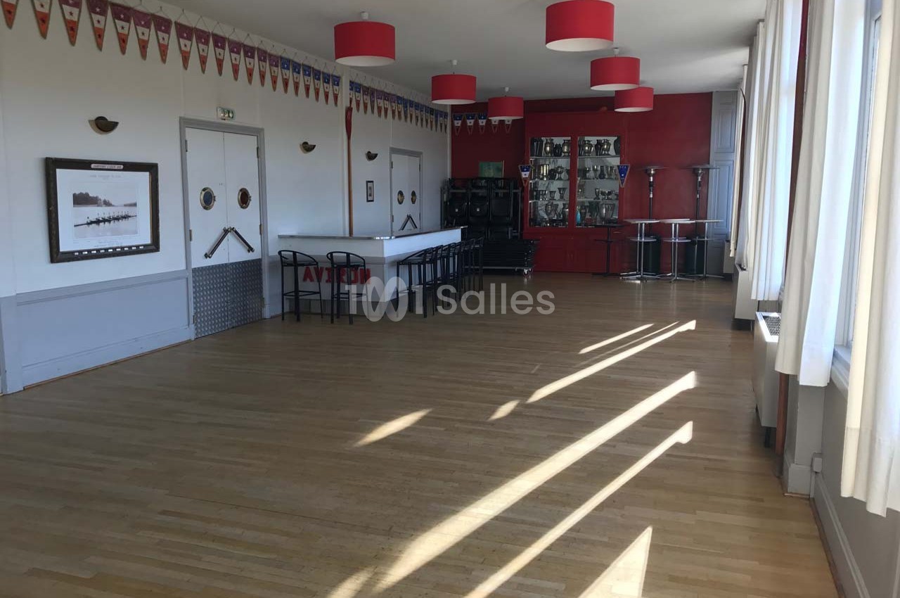 Salle lumineuse avec parquet en bois, bar rouge, chaises noires, décorations murales et grandes fenêtres laissant entrer la…