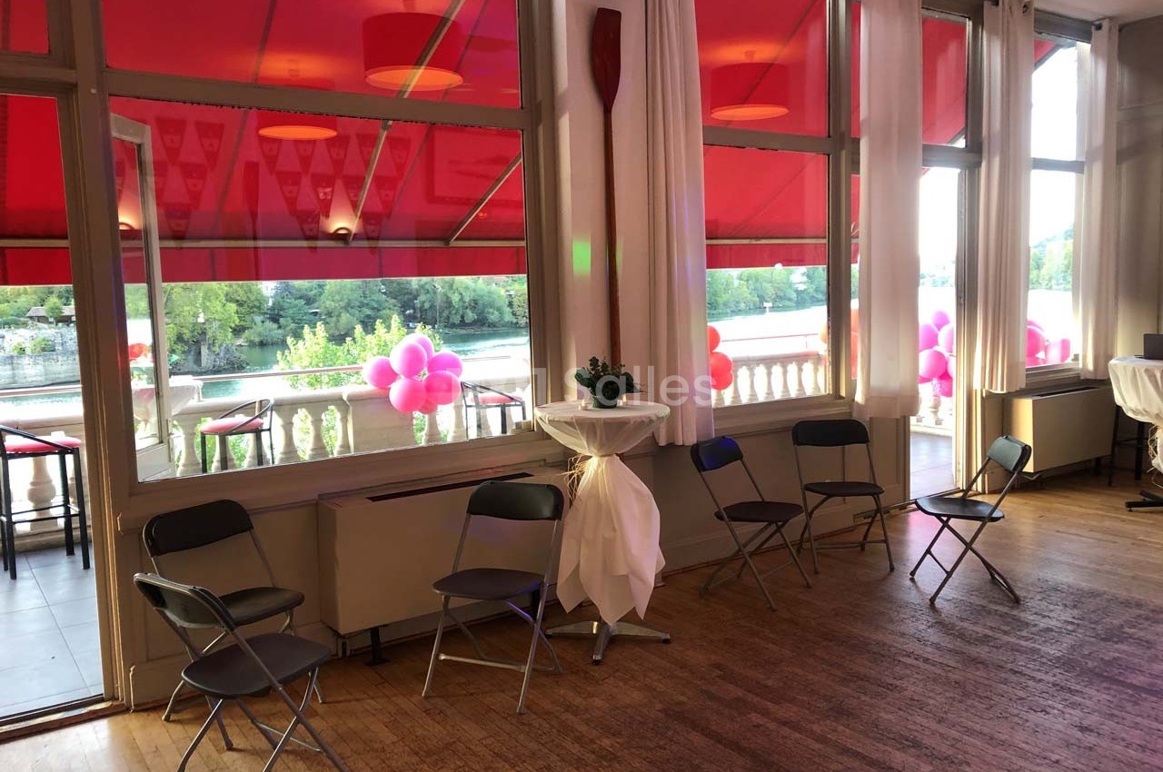 Salle lumineuse avec parquet, chaises pliantes, ballons roses en extérieur et vue sur une rivière bordée d'arbres.