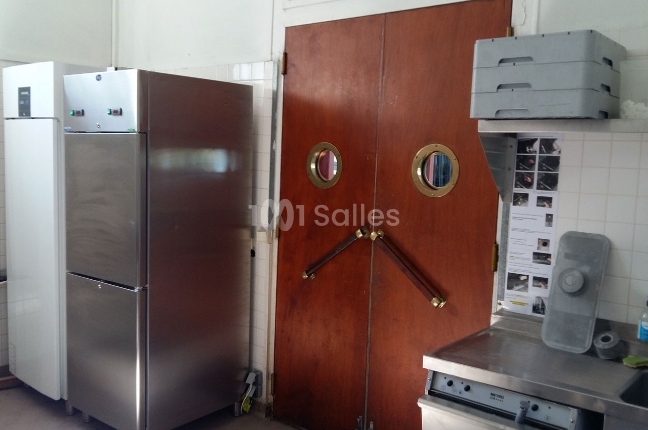 Cuisine professionnelle avec réfrigérateur en acier inoxydable, portes battantes en bois et plans de travail en métal.