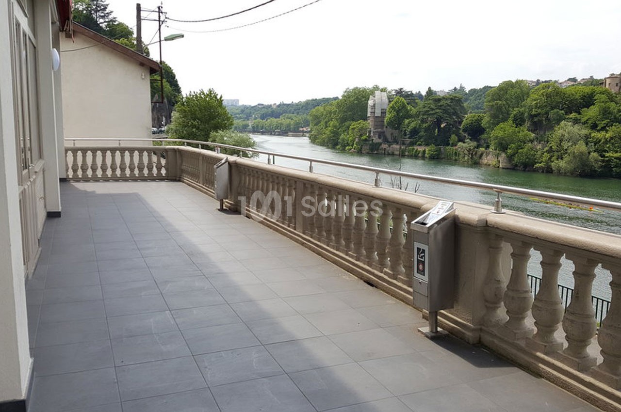 Terrasse carrelée avec balustrade donnant sur une rivière bordée de végétation et bâtiments en arrière-plan.