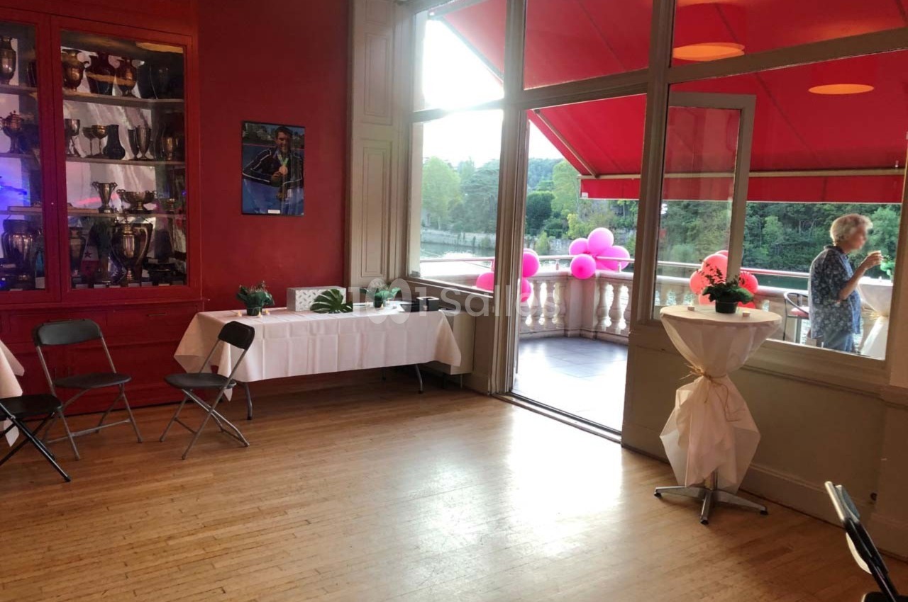 Salle lumineuse avec parquet, tables décorées, baies vitrées ouvertes sur une terrasse avec vue sur un espace verdoyant.