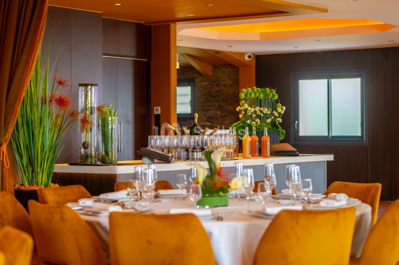 Salle de restaurant élégante avec table dressée, chaises jaunes et bar en arrière-plan décoré de plantes et bouteilles.