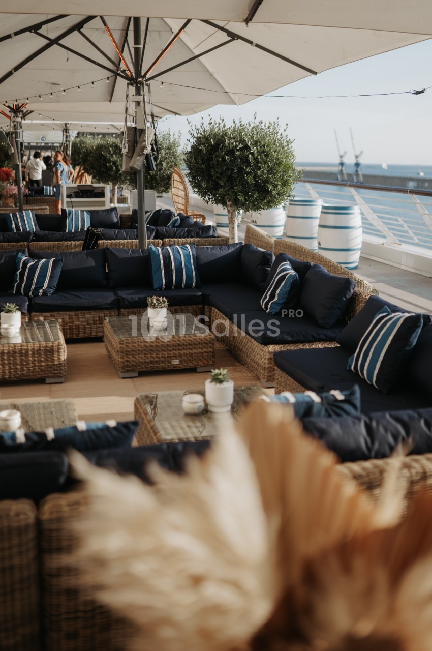 Terrasse aménagée avec des canapés en rotin, coussins bleus, tables basses et vue sur la mer en arrière-plan.