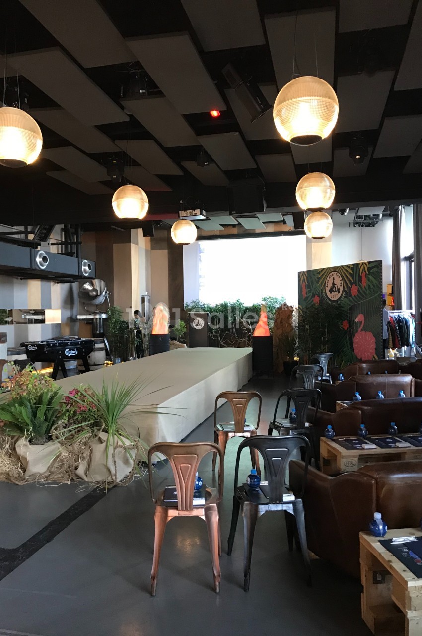 Salle aménagée avec des chaises, des tables, une scène, des plantes décoratives et des suspensions lumineuses.