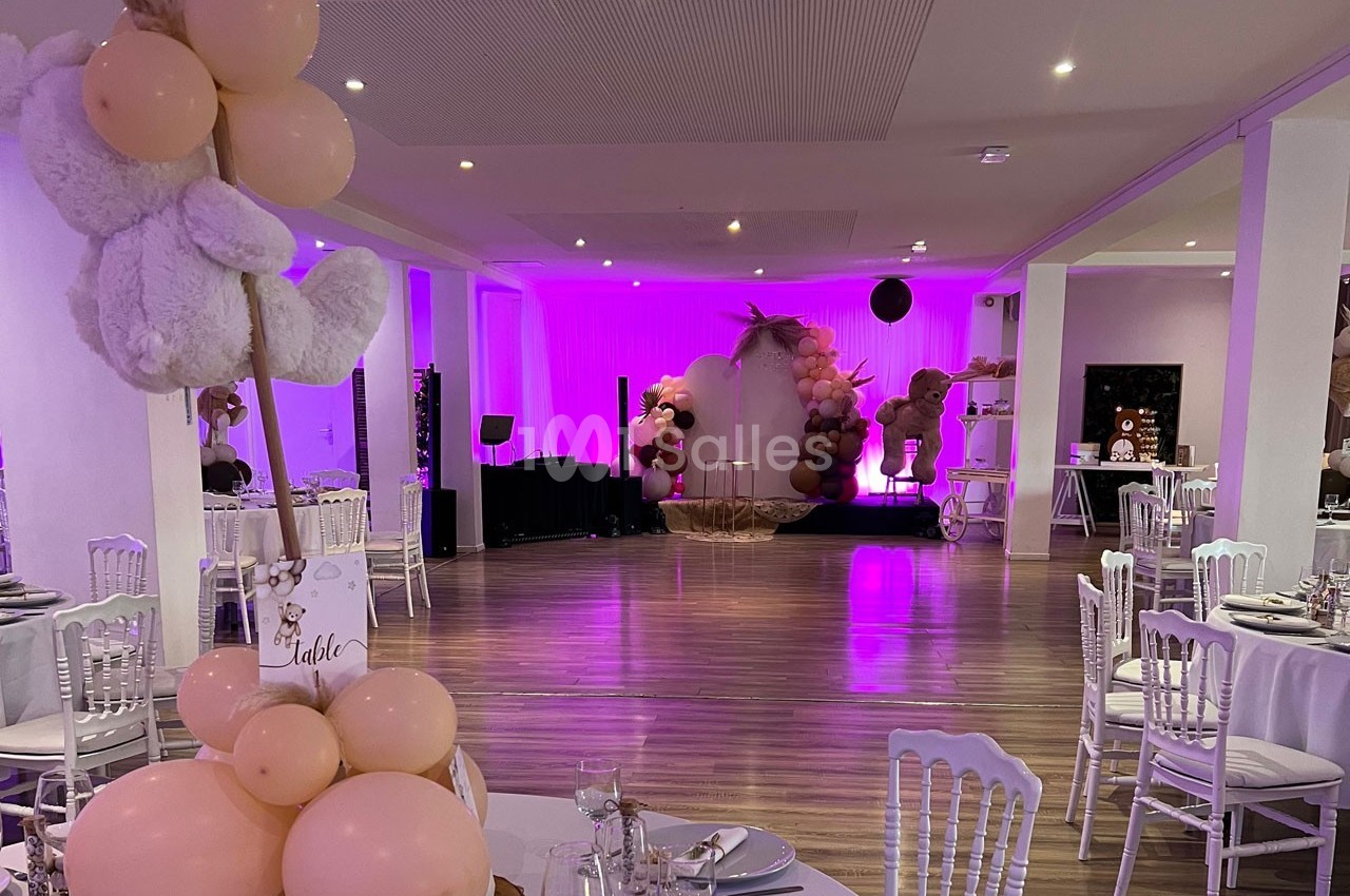 Salle de réception décorée avec des ballons roses et beiges, tables dressées et éclairage violet en arrière-plan.