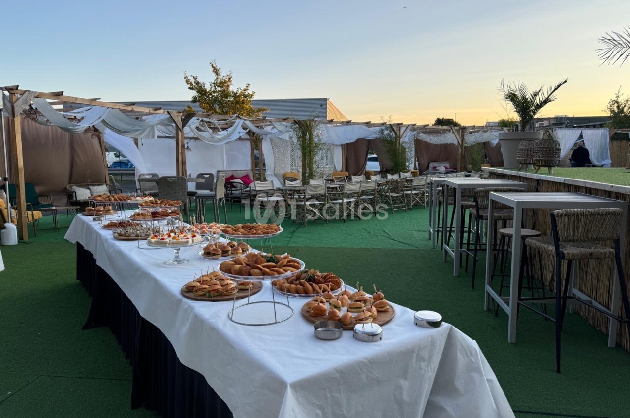 Buffet en extérieur avec tables dressées, chaises et décoration blanche sous un ciel au coucher du soleil.