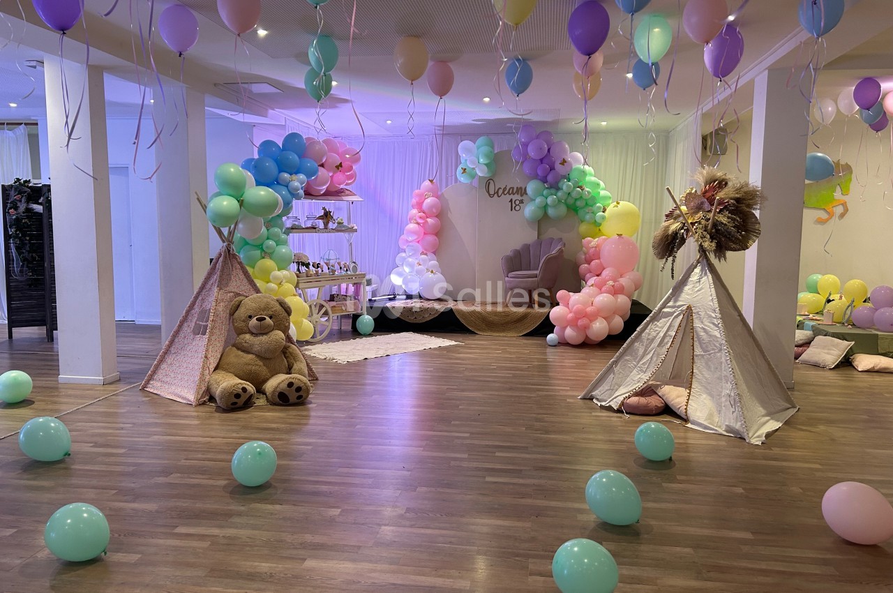 Salle décorée pour une fête avec ballons colorés, tipis, un grand ours en peluche et une arche de ballons pastel.