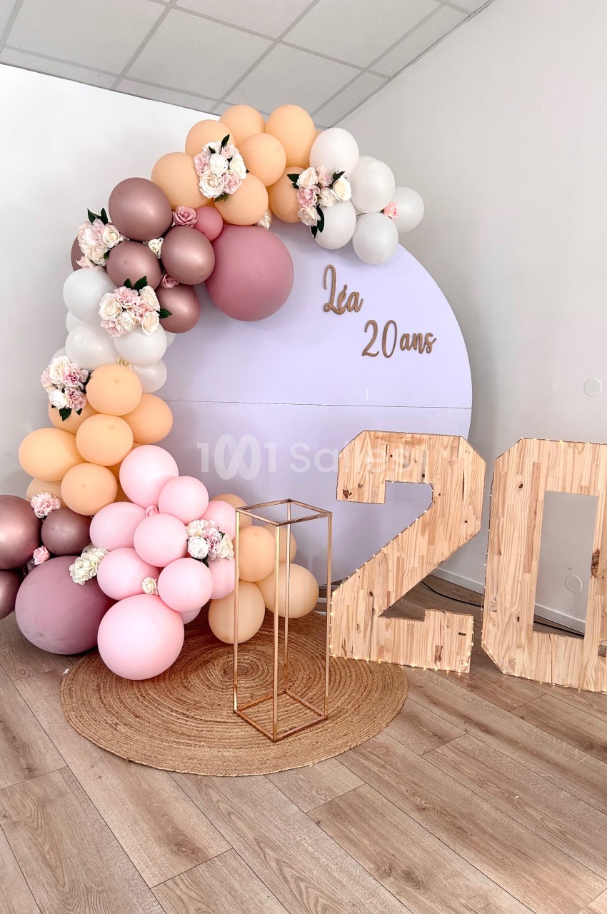 Décoration d'anniversaire avec arche de ballons pastel, panneau ’Léa 20 ans’ et chiffres en bois sur un tapis rond.