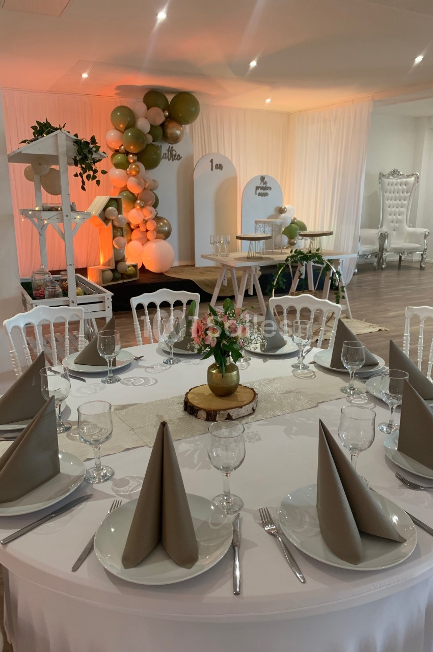 Salle décorée pour un événement avec ballons, table dressée, chaises blanches et éclairage chaleureux.