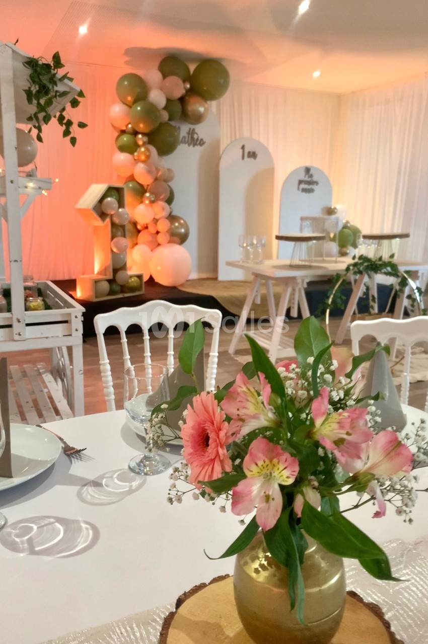 Salle décorée pour un événement, avec ballons, fleurs sur table et mobilier blanc dans une ambiance lumineuse.
