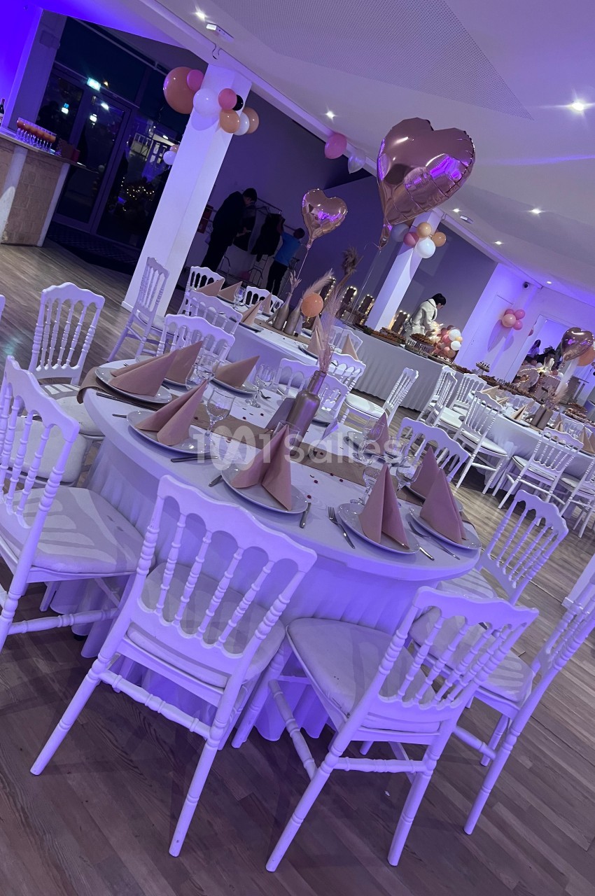 Salle décorée pour un événement avec tables rondes, chaises blanches, serviettes pliées et ballons roses en arrière-plan.