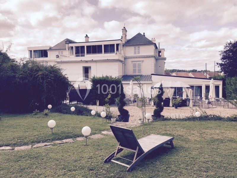 Façade d'une grande maison blanche avec jardin, terrasse aménagée et transat au premier plan.
