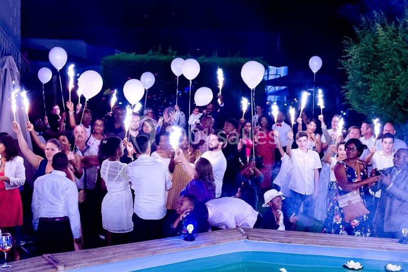 Groupe de personnes célébrant en soirée autour d'une piscine, tenant des ballons et des cierges magiques allumés.