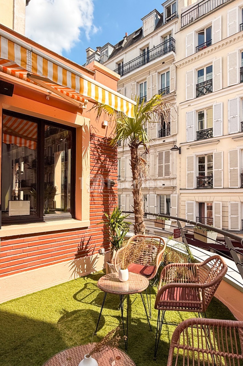 Terrasse ensoleillée avec mobilier en rotin, plantes et vue sur des immeubles parisiens aux façades claires.