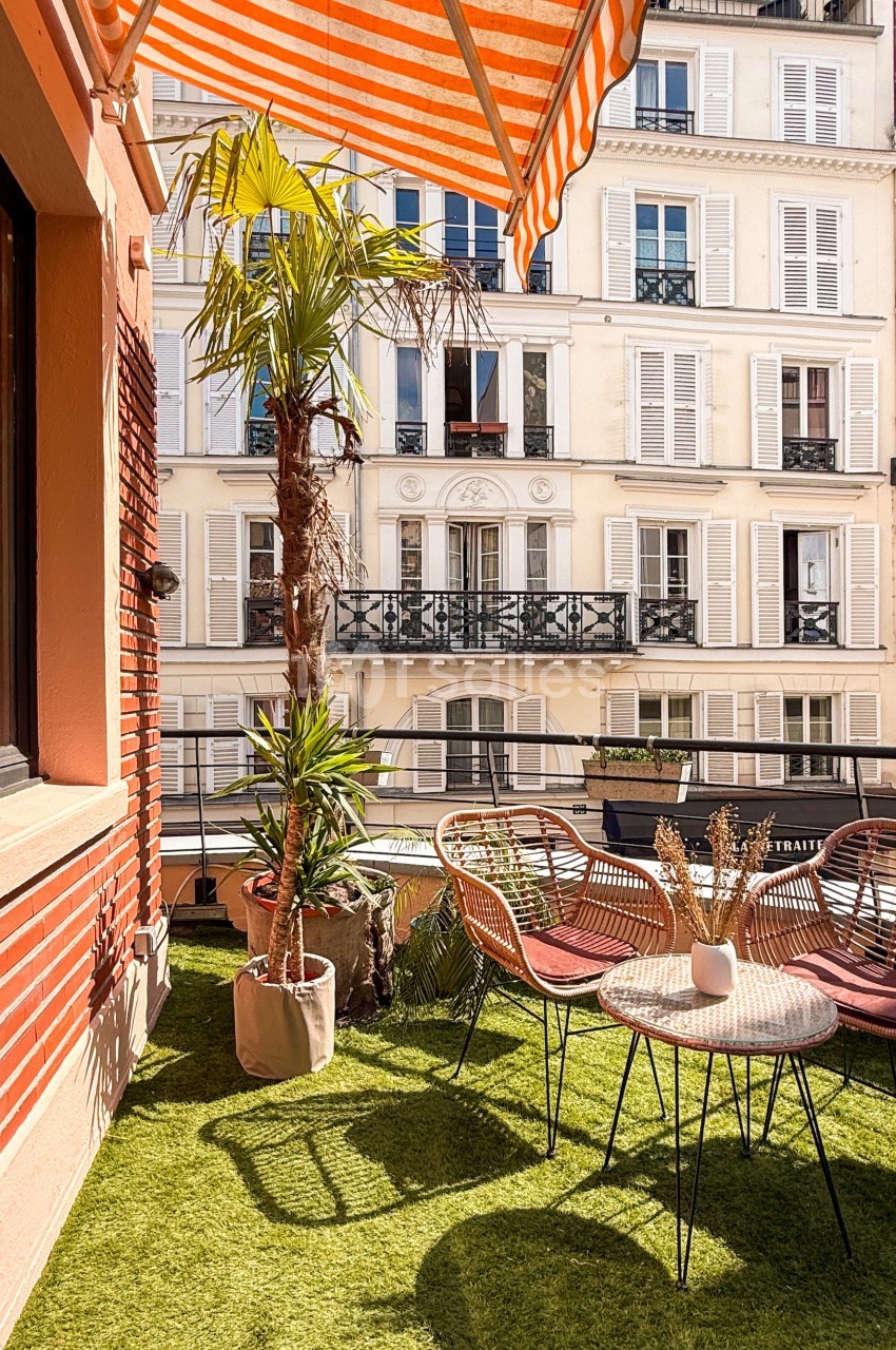 Terrasse avec gazon artificiel, mobilier en rotin, plantes et vue sur des immeubles parisiens aux volets blancs.