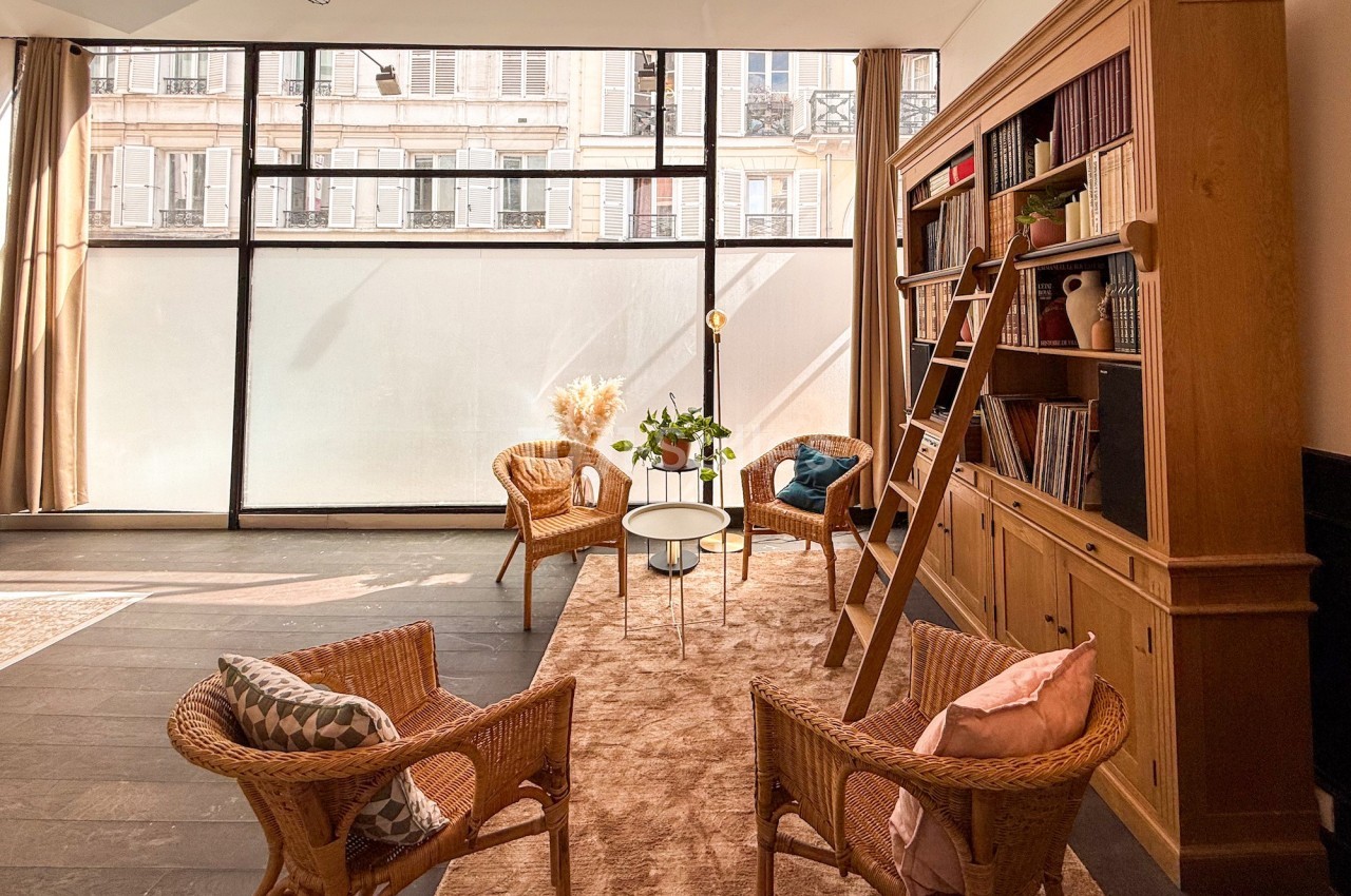 Salon lumineux avec grandes baies vitrées, bibliothèque en bois, fauteuils en rotin et table basse sur tapis beige.