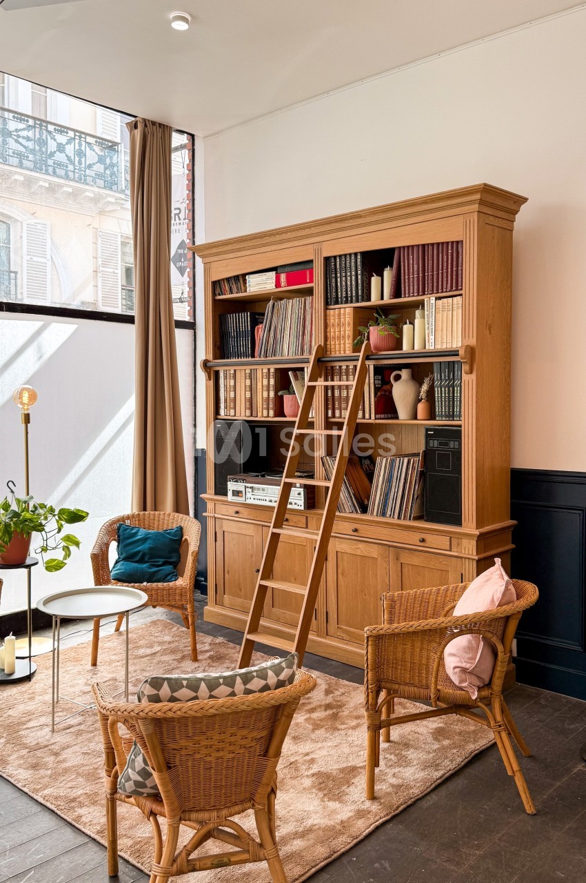 Bibliothèque en bois avec échelle, entourée de fauteuils en rotin et tables basses dans un salon lumineux.