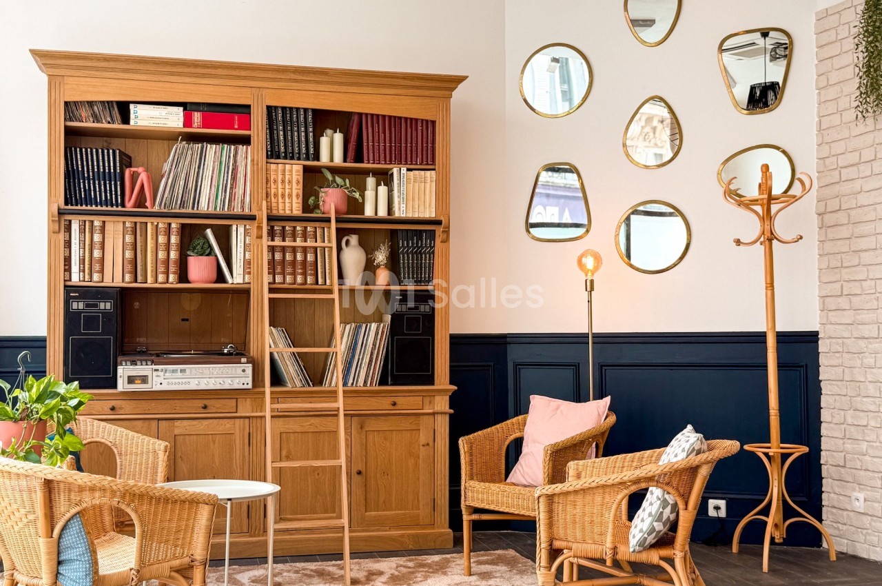 Bibliothèque en bois avec livres et vinyles, fauteuils en rotin, lampadaire et miroirs décoratifs sur un mur blanc.
