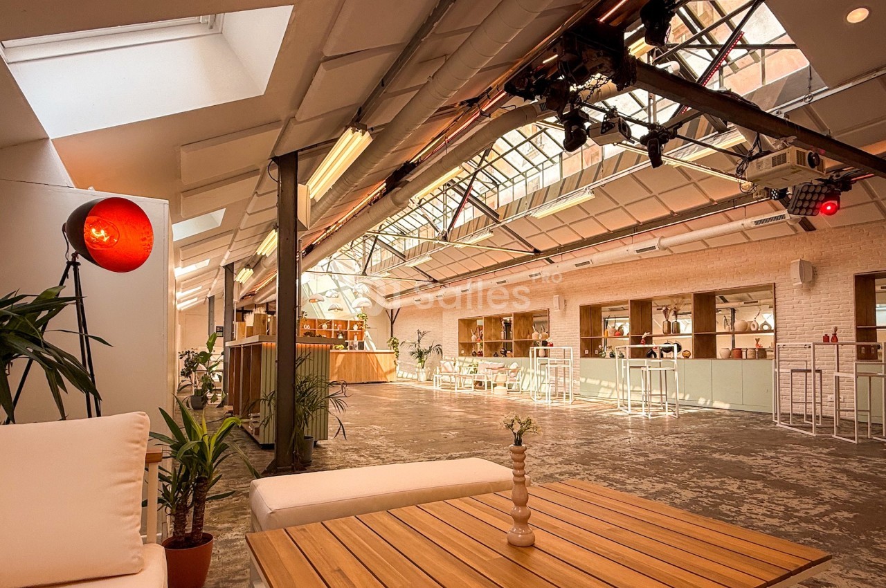 Espace lumineux avec verrière, mobilier en bois, plantes et éclairage chaleureux dans un cadre industriel.