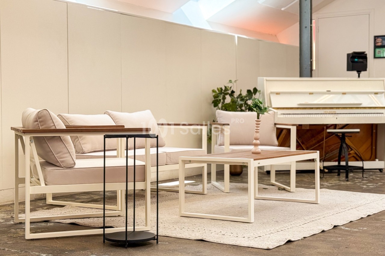 Salon lumineux avec canapé beige, table basse blanche, tapis clair, petite table noire et piano en arrière-plan.