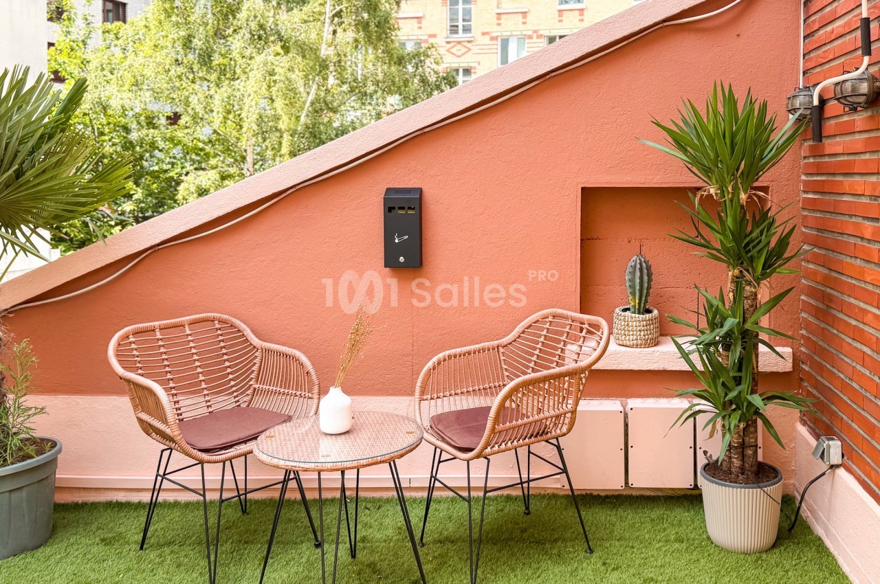 Terrasse avec table ronde, deux fauteuils en rotin, plantes en pot et mur rose, sur fond de verdure et briques.