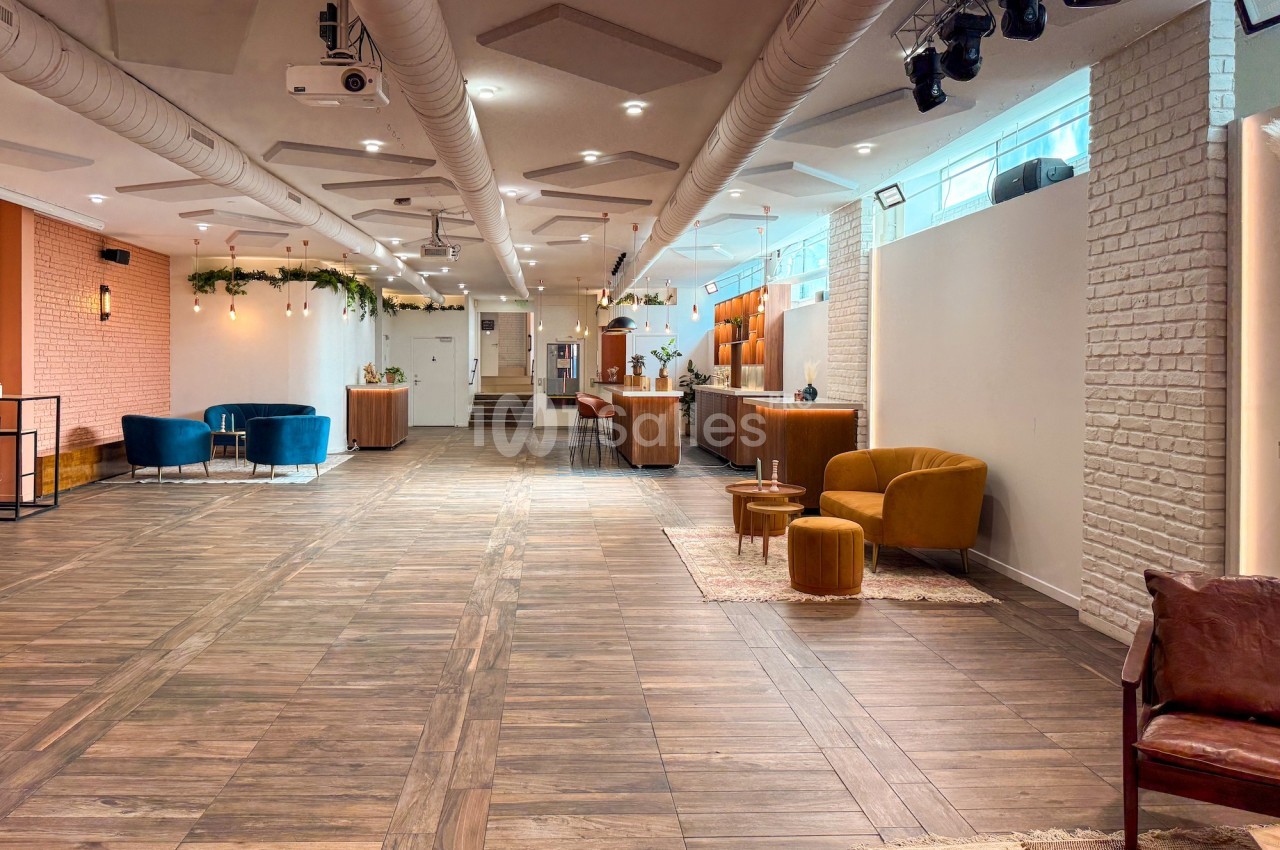 Salle lumineuse avec parquet, fauteuils colorés, murs en briques et éclairage suspendu, aménagée pour des événements.