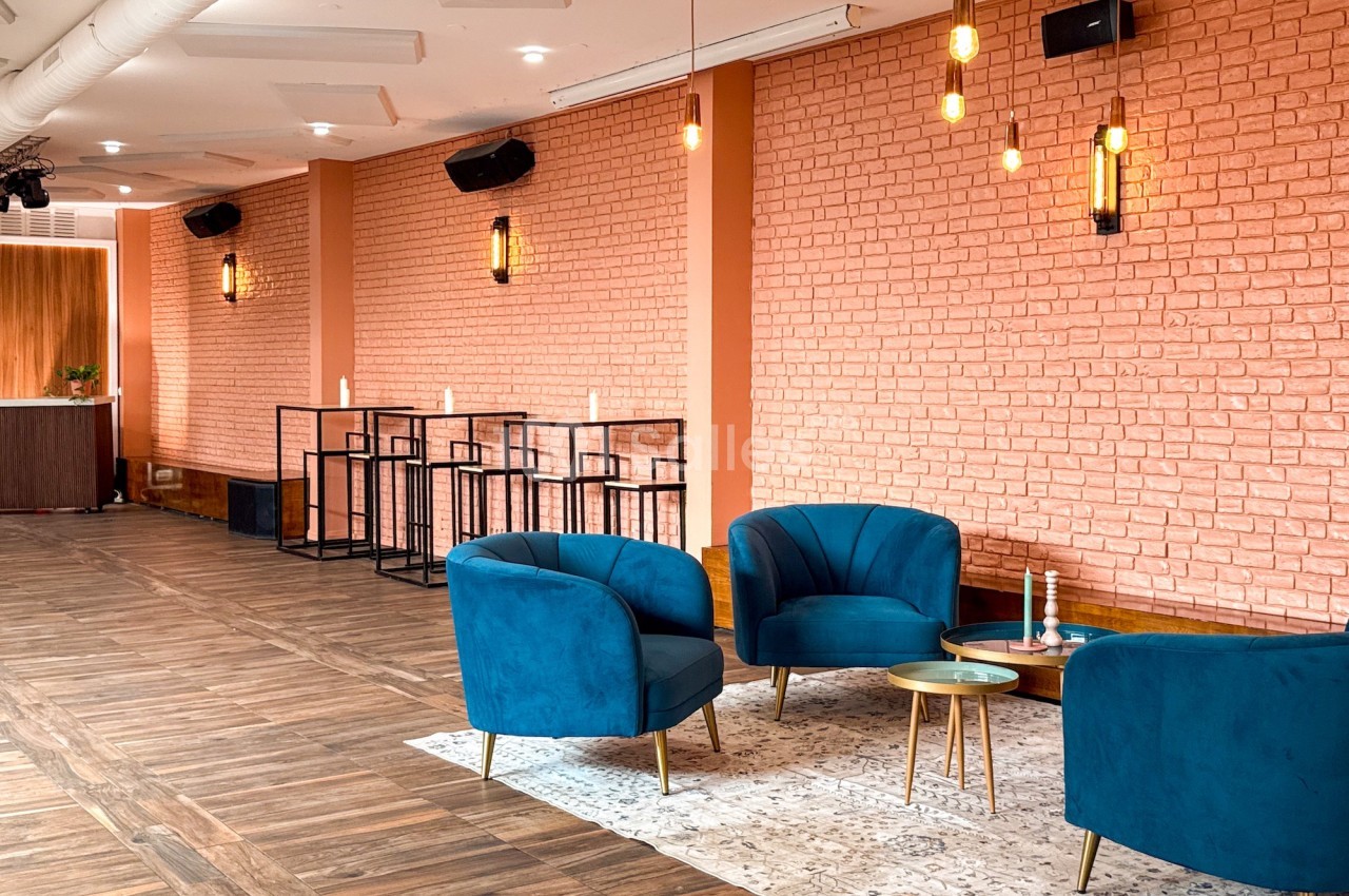 Salle lumineuse avec murs en briques, fauteuils bleus, tables basses dorées et éclairage suspendu moderne.