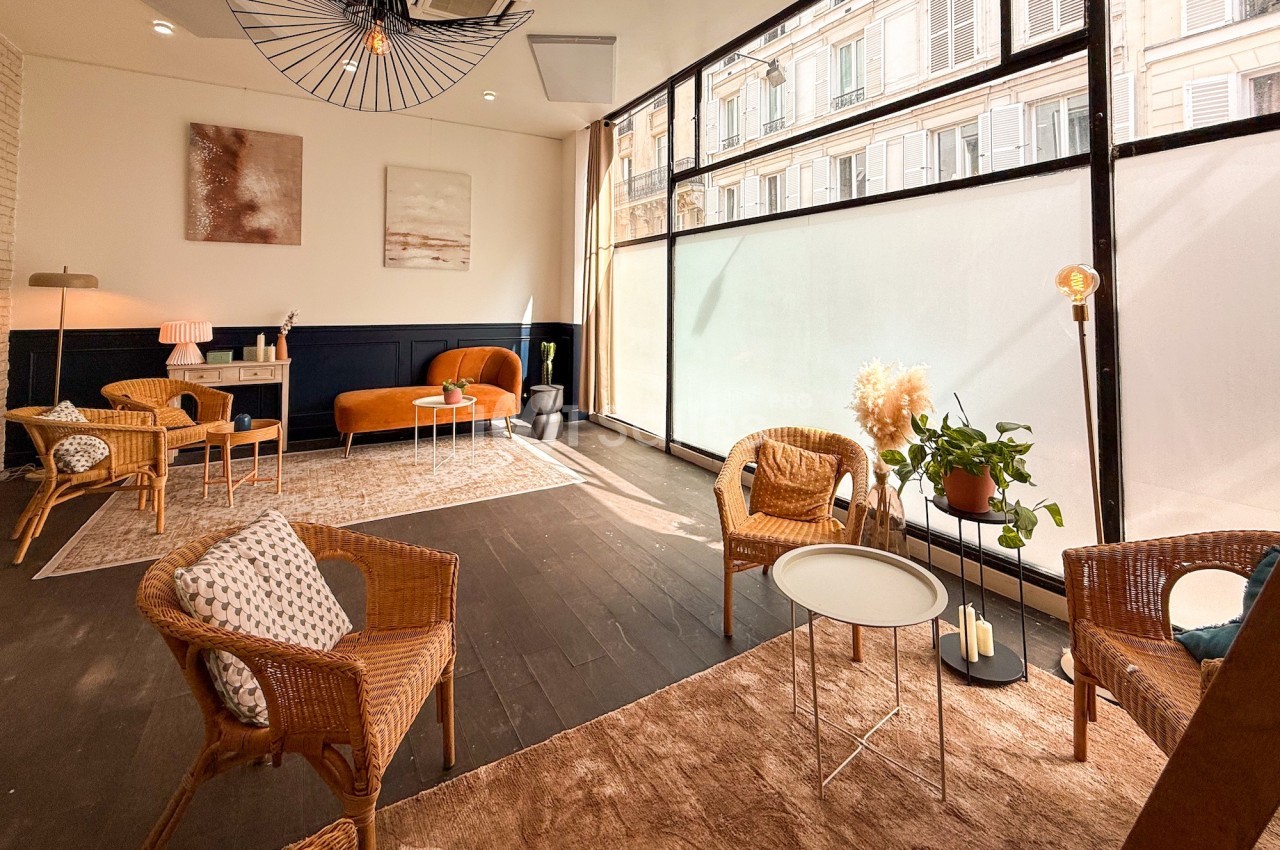 Espace lumineux avec fauteuils en rotin, canapé orange, plantes et grandes baies vitrées donnant sur l'extérieur.